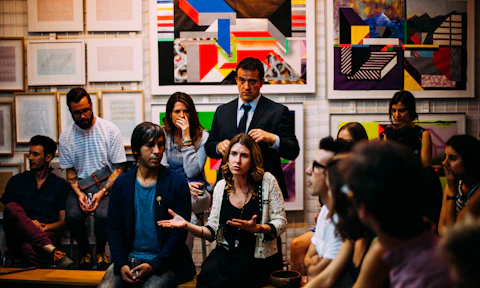 A woman speaks to a seated group in a room hung with colorful art.