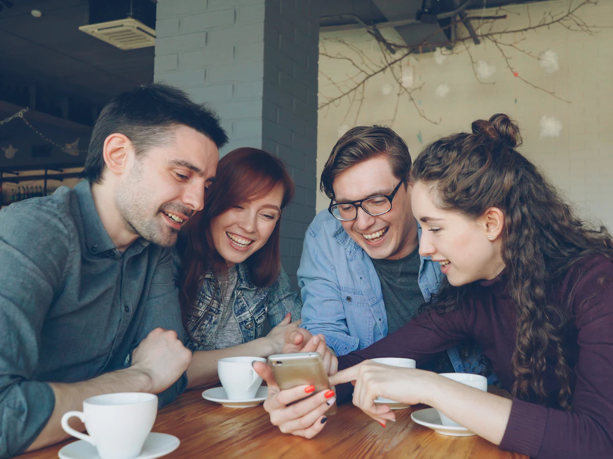 Four friends at a cafe table look at one phone together and laugh.